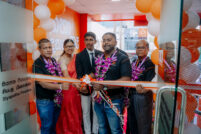 Group of six people at a ribbon-cutting ceremony inside an orange-themed storefront, with balloons and floral leis visible in the scene.